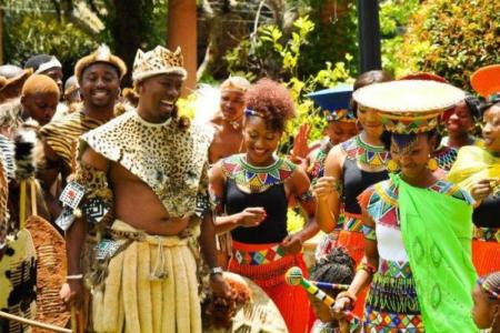 A group of people in traditional Zulu cultural attire laugh and dance against a green leafy background.