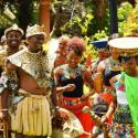 A group of people in traditional Zulu cultural attire laugh and dance against a green leafy background.